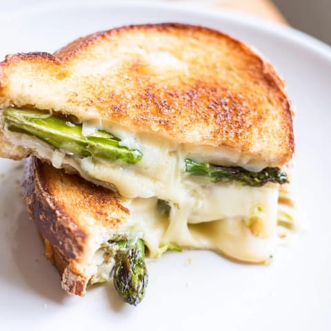 A close-up of Asparagus & Brie Grilled Cheese shows oozing cheese and tender green spears between crispy toasted bread.