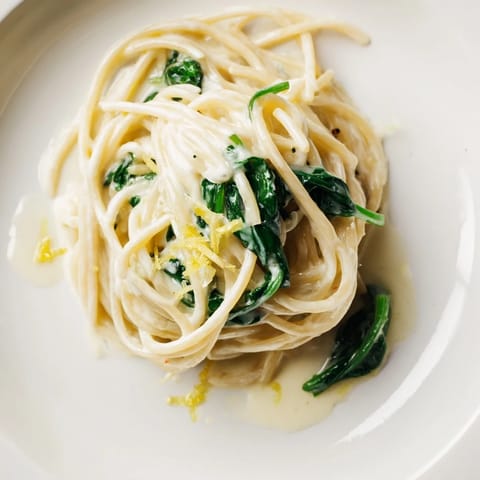 A close-up of silky lemon cream sauce coating al dente spaghetti and vibrant wilted spinach leaves.  